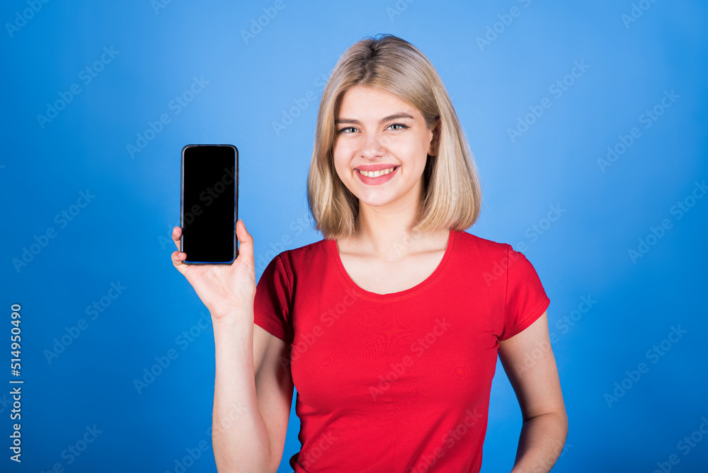 Young and attractive caucasian blonde girl in casual clothes showing the screen of a mobile phone with copy space for advertising isolated on a blue studio background. Young and attractive caucasian blonde girl in casual clothes showing the screen of a mobile phone with copy space for advertising isolated on a blue studio background.