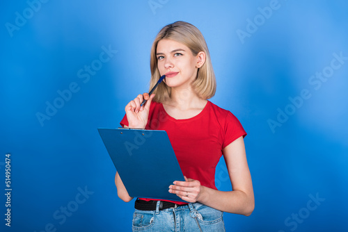 A young and attractive Caucasian blonde girl in casual clothes holding a clipboard and a pen near her mouth and thinking isolated on a blue studio background.
