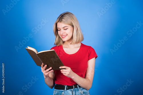Young and attractive caucasian blonde girl in casual clothes smiling, holding and reading a book isolated on a blue studio background. Education concept.