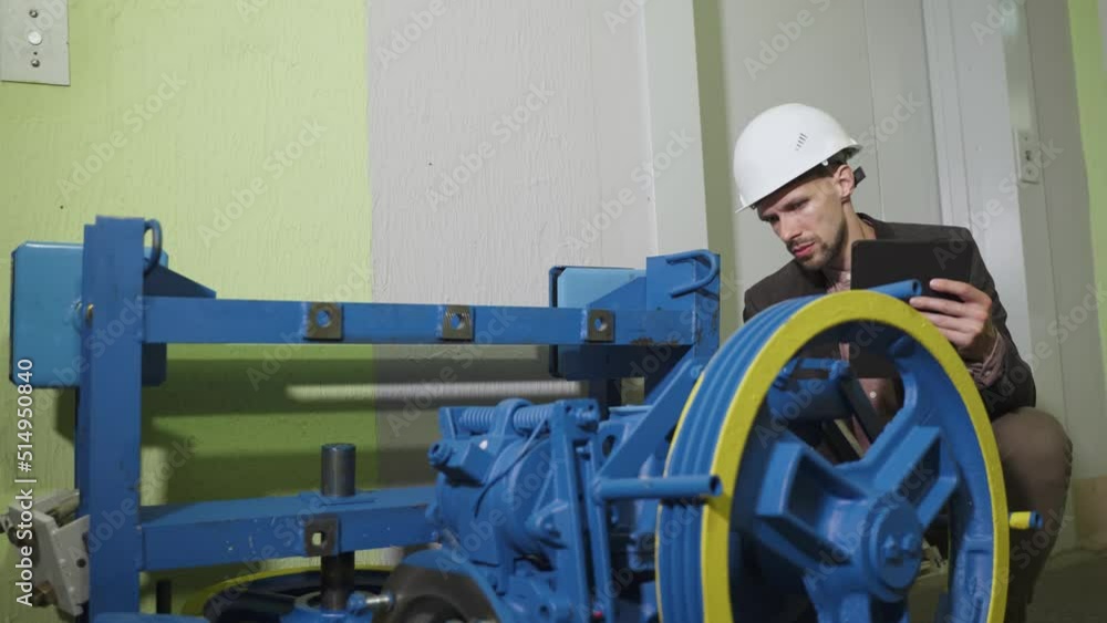Controller in helmet concentrated, carefully checks technical parameters, quality of new elevator equipment, comparing it with data in tablet. Foreman evaluates and accepts the electric drive. Controller in helmet concentrated, carefully checks technical parameters, quality of new elevator equipment, comparing it with data in tablet. Foreman evaluates and accepts the electric drive.