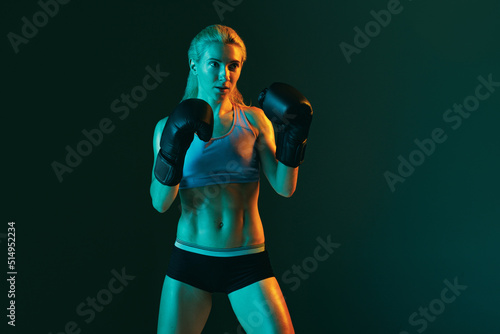 One young woman, female professional kickboxer in sports uniform and gloves training isolated on dark green background in neon.