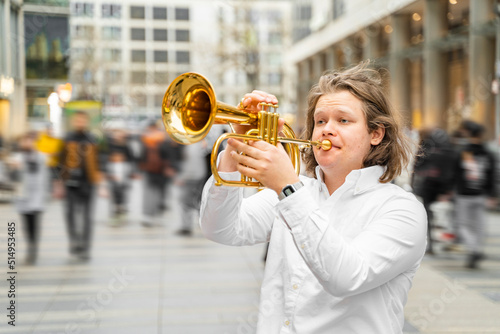 Young caucasian long-haired blond man in white shirt playing jazz on golden trumpet with pleasure standing in the middle of crowded downtown street with many people and department stores. Hobby