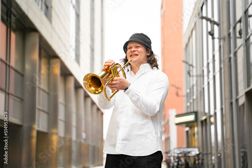 Young caucasian blond cheerful smiling man in white shirt and hat playing funky jazz on golden trumpet with pleasure standing between modern skyscrapers in downtown with department stores, in Germany