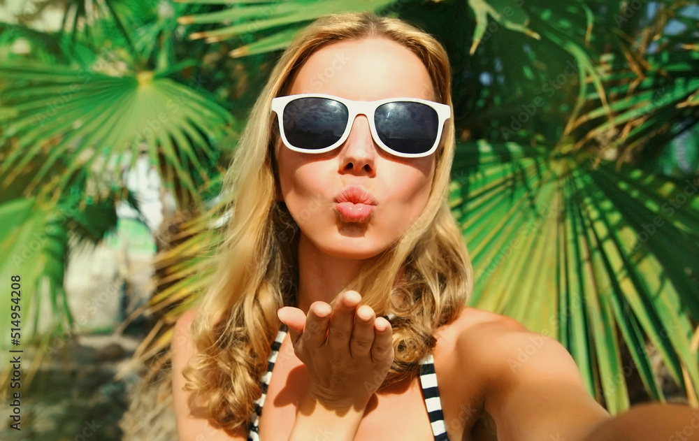 Summer portrait of young woman stretching her hand for taking selfie with smartphone blowing her lips sending sweet air kiss on palm tree background