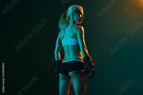 Slika na platnu Back view of young woman, female professional kickboxer in sports uniform and gloves training isolated on dark green background in neon