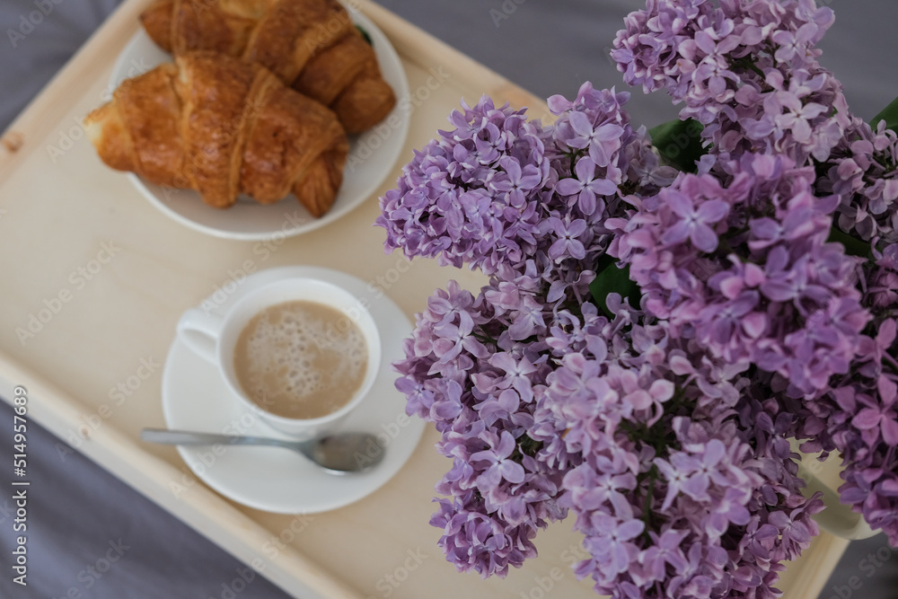 Breakfast in bed. Tray with coffee, croissants and a bouquet of lilacs on the bed in the bedroom Breakfast in bed. Tray with coffee, croissants and a bouquet of lilacs on the bed in the bedroom