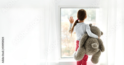 Loneliness. Lonely orphan girl in an orphanage looking outside while holding teddy bear the window. Child feelings concept photo