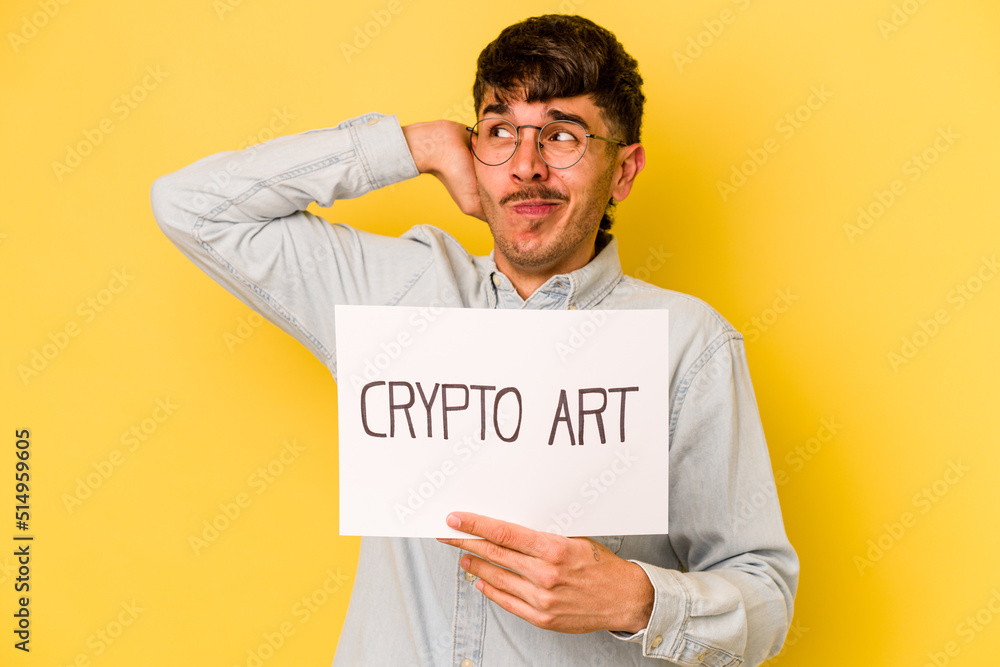 Young hispanic man holding crypto art placard isolated on yellow background touching back of head, thinking and making a choice. Young hispanic man holding crypto art placard isolated on yellow background touching back of head, thinking and making a choice.