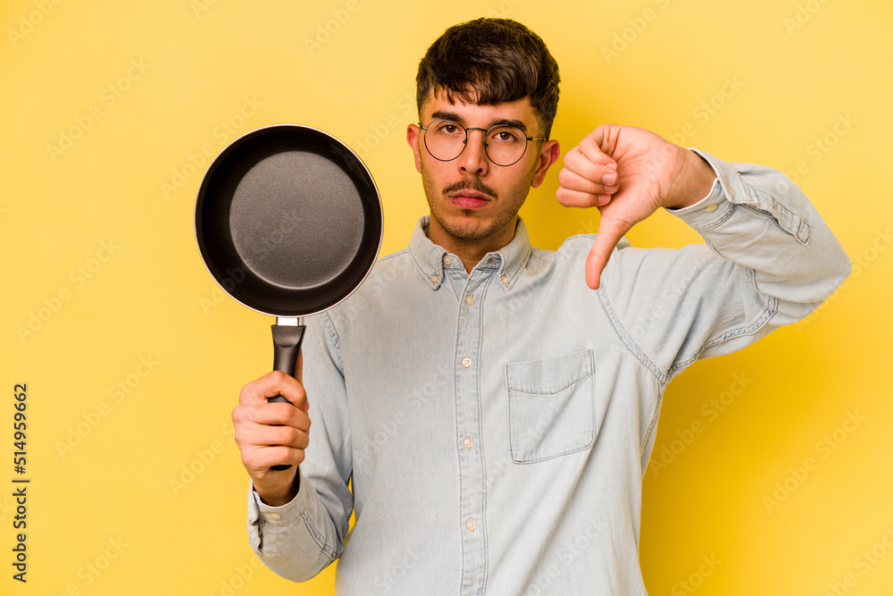 Young hispanic cooker holding frying pan isolated on yellow background showing a dislike gesture, thumbs down. Disagreement concept. Young hispanic cooker holding frying pan isolated on yellow background showing a dislike gesture, thumbs down. Disagreement concept.
