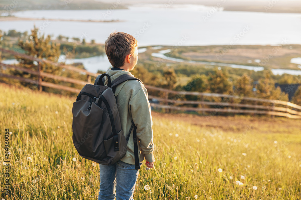 Tourist with backpack standing on top of hill in grass field and enjoying beautiful landscape view. Rear view of teenage boy hiker resting in nature. Active lifestyle. Concept of local travel Tourist with backpack standing on top of hill in grass field and enjoying beautiful landscape view. Rear view of teenage boy hiker resting in nature. Active lifestyle. Concept of local travel