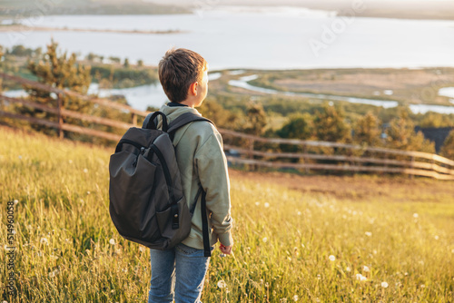 Tourist with backpack standing on top of hill in grass field and enjoying beautiful landscape view. Rear view of teenage boy hiker resting in nature. Active lifestyle. Concept of local travel © Lyubov