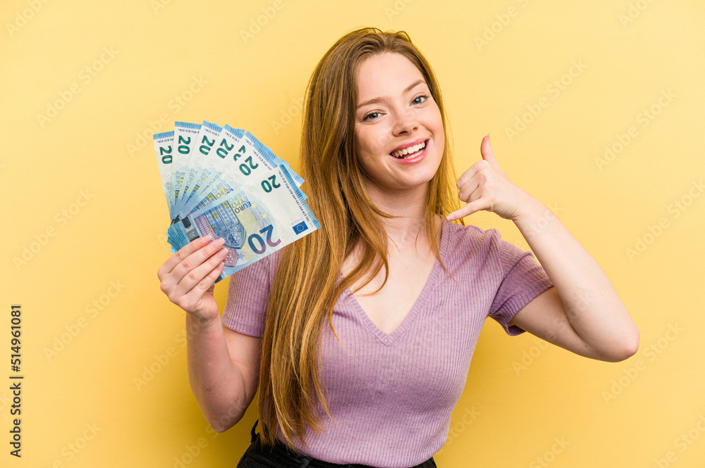 Young caucasian woman holding banknotes isolated on yellow background showing a mobile phone call gesture with fingers. Young caucasian woman holding banknotes isolated on yellow background showing a mobile phone call gesture with fingers.