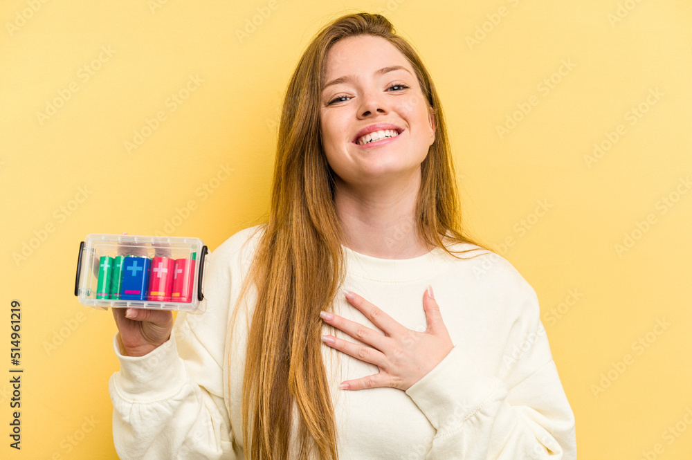 Young caucasian woman holding a battery box isolated on yellow background laughs out loudly keeping hand on chest. Young caucasian woman holding a battery box isolated on yellow background laughs out loudly keeping hand on chest.