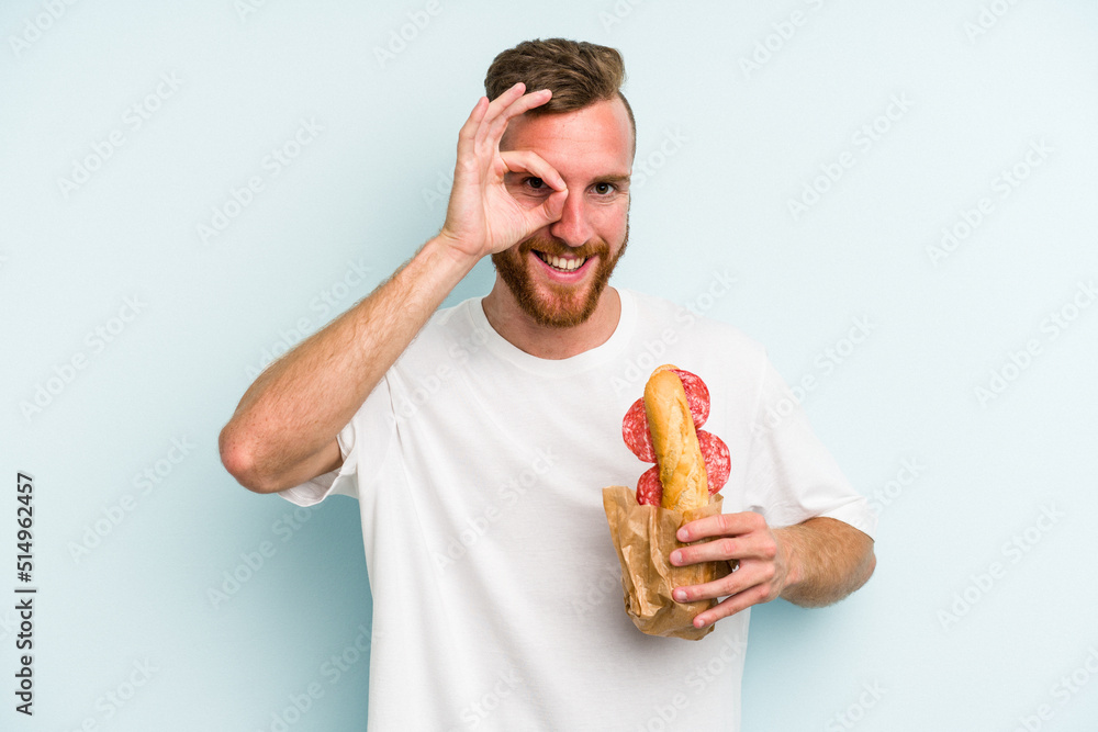 Young caucasian man eating a sandwich isolated on blue background excited keeping ok gesture on eye. Young caucasian man eating a sandwich isolated on blue background excited keeping ok gesture on eye.