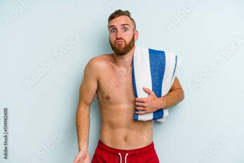 Young caucasian man going to the beach holding a towel isolated on blue background shrugs shoulders and open eyes confused. photo