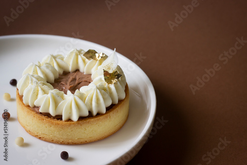 Mini tart with chocolate, edible gold and whipped cream. White plate, brown background