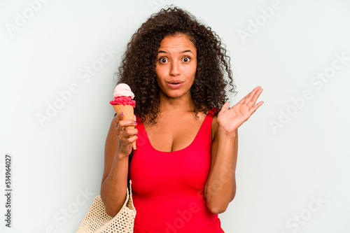 Young brazilian woman going to the beach holding an ice cream isolated on blue background surprised and shocked.