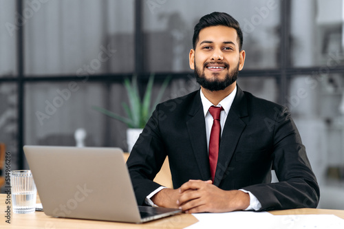 Portrait of a positive young male confident arabian or indian businessman, successful entrepreneur in a suit, sit at a work desk with laptop in modern creative office, looks at camera, smile friendly © Kateryna