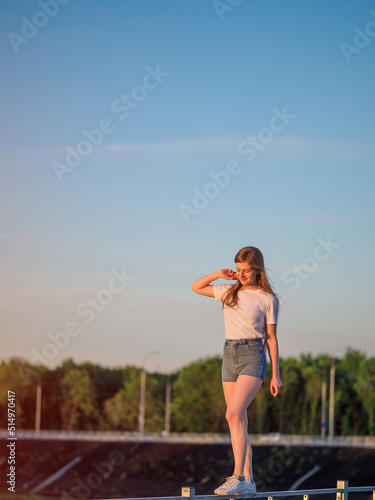 A young beautiful woman in a white blouse and shorts walks along the railing against the sky, keeping her balance. Happy Summer time