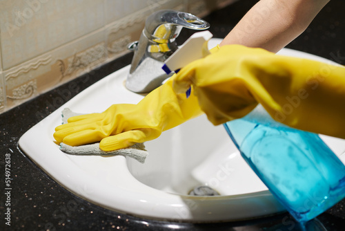 Close-up of woman in protective rubber gloves washing sink with rag and spray in bathroom