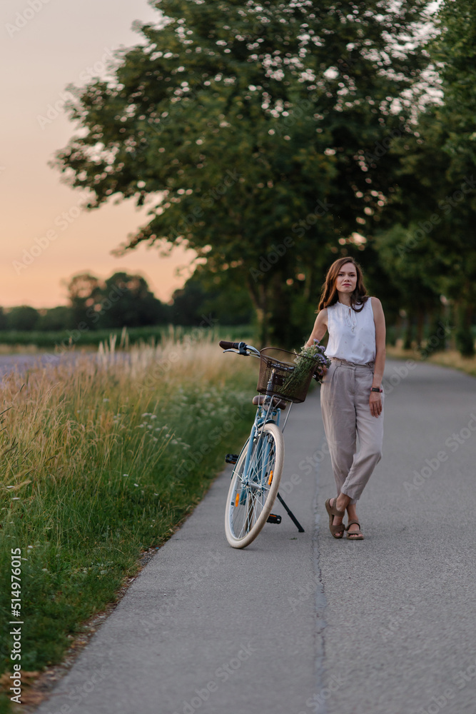 Romantic portrait of beautiful woman. A girl with a bouquet and a blue bicycle in countryside. Summertime careless and happiness. Walk in nature. Beautiful sunset. purple field. Meadow. Freedom mood
