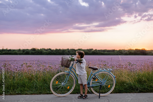 Romantic portrait of an adorable child. A boy with a bouquet and a bicycle in countryside. Summertime childhood and happiness. Walk in nature. Beautiful sunset. School holidays. purple field. Meadow photo