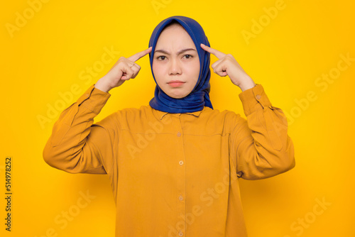 Pensive young Asian Muslim woman dressed in orange shirt trying to remember something isolated on yellow background © Sewupari Studio