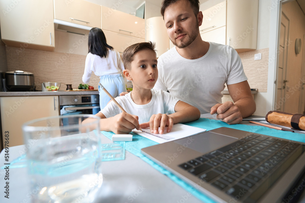 Man helping child with homework at laptop in kitchen Man helping child with homework at laptop in kitchen