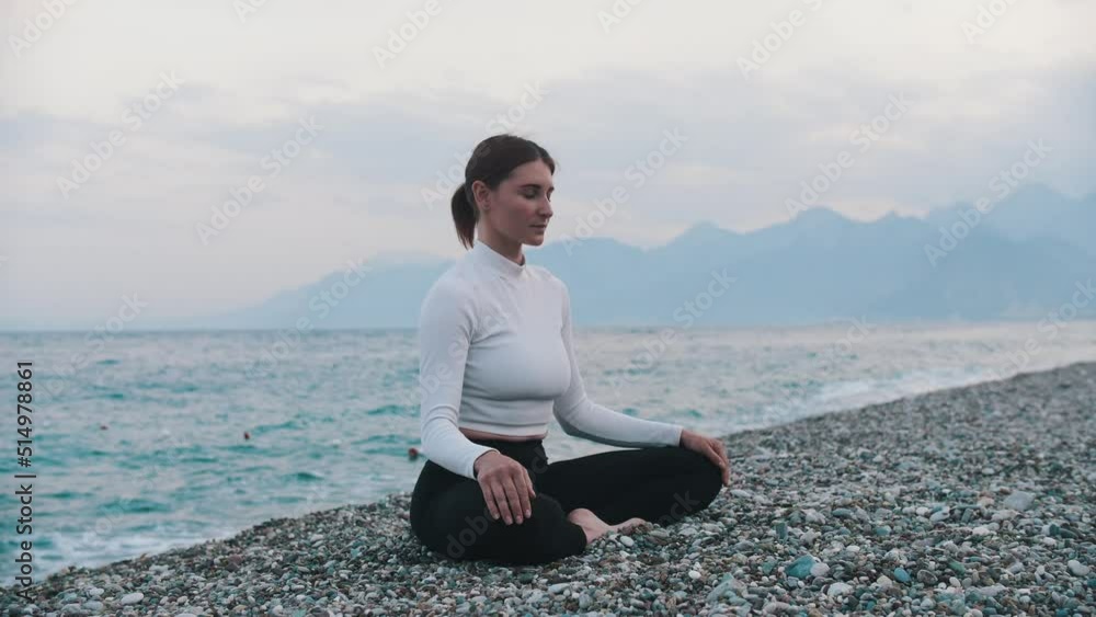 A woman in white sweater meditating on the pebble beach by the blue sea A woman in white sweater meditating on the pebble beach by the blue sea