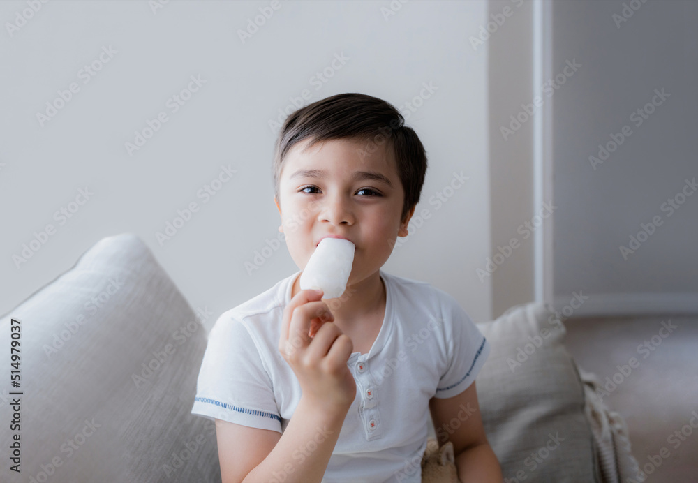 Happy kid eating ice lolly, Portrait of cute little boy sitting on sofa next to a window having a refreshment, Child with a smiling face relaxing at home. Kid looking at camera while eating ice cream Happy kid eating ice lolly, Portrait of cute little boy sitting on sofa next to a window having a refreshment, Child with a smiling face relaxing at home. Kid looking at camera while eating ice cream