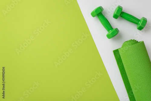 Fitness accessories concept. Top view photo of green sports mat and dumbbells on bicolor green and white background with copyspace photo