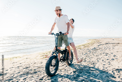 Cheerful friends couple on bicycle having fun with electric bike - carefree boys having fun and smiling on bicycle on the beach on a sunny day - freedom concept