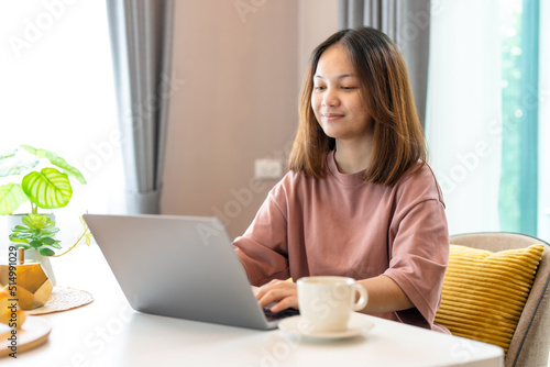 young Asian woman with long hair sitting in the living room on the sofa using a laptop to video call a coworker. During the conversation, typed a message as well. Work from home.