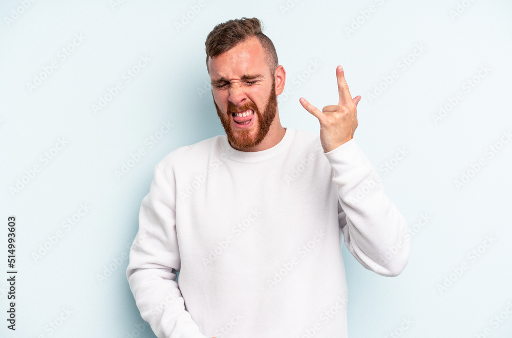 Young caucasian man isolated on blue background showing rock gesture with fingers Young caucasian man isolated on blue background showing rock gesture with fingers