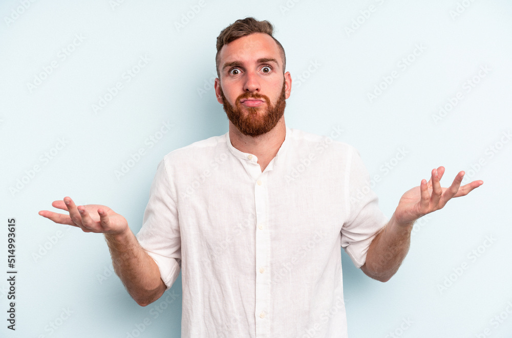 Young caucasian man isolated on blue background doubting and shrugging shoulders in questioning gesture. Young caucasian man isolated on blue background doubting and shrugging shoulders in questioning gesture.
