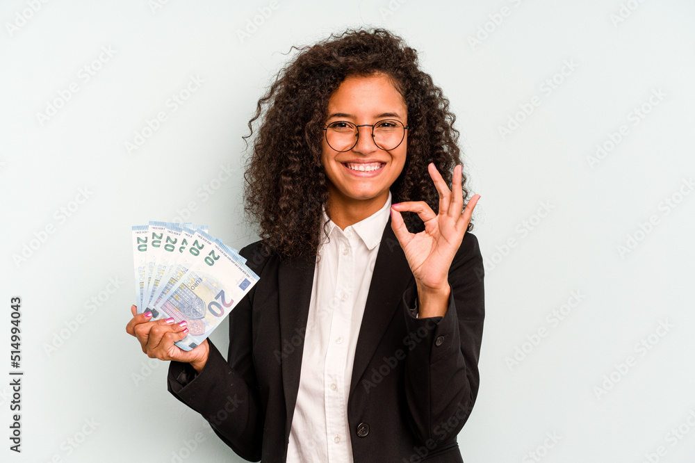 Young business Brazilian woman holding banknotes isolated on white background cheerful and confident showing ok gesture. Young business Brazilian woman holding banknotes isolated on white background cheerful and confident showing ok gesture.
