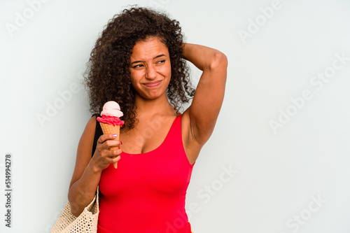 Young brazilian woman going to the beach holding an ice cream isolated on blue background touching back of head, thinking and making a choice.