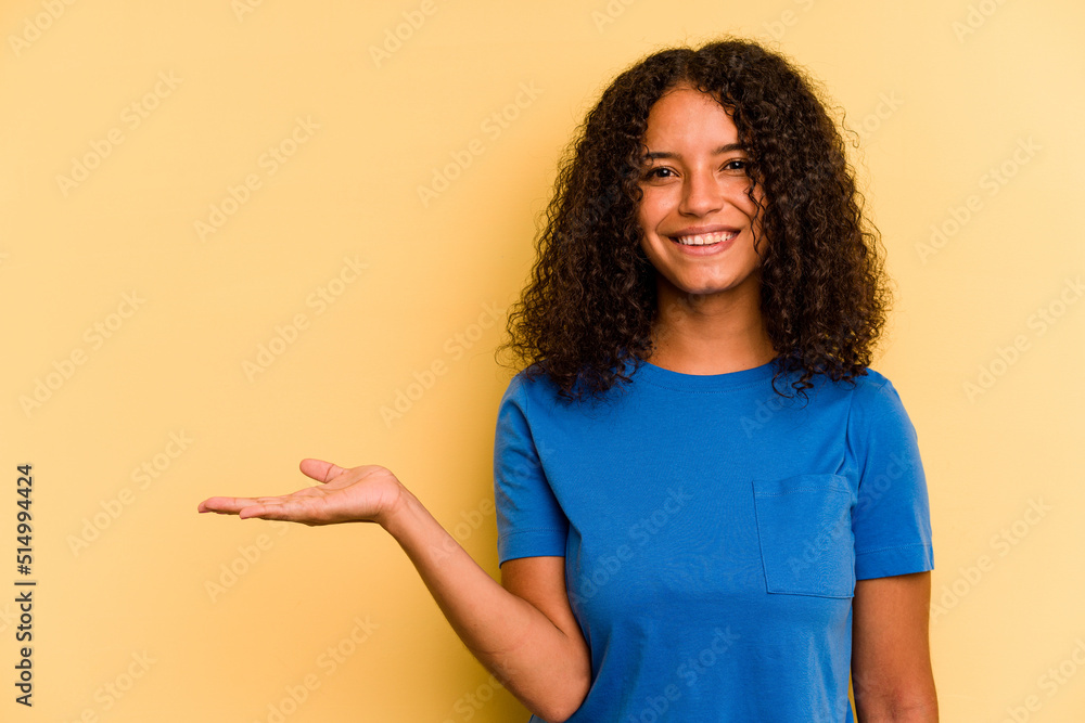 Young Brazilian woman isolated on yellow background showing a copy space on a palm and holding another hand on waist. Young Brazilian woman isolated on yellow background showing a copy space on a palm and holding another hand on waist.