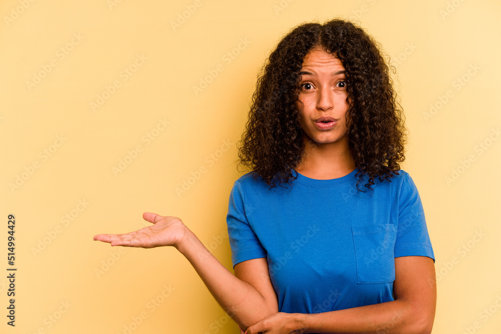 Young Brazilian woman isolated on yellow background impressed holding copy space on palm. Young Brazilian woman isolated on yellow background impressed holding copy space on palm.