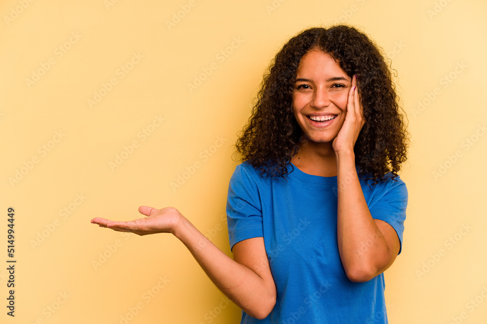Young Brazilian woman isolated on yellow background holds copy space on a palm, keep hand over cheek. Amazed and delighted. Young Brazilian woman isolated on yellow background holds copy space on a palm, keep hand over cheek. Amazed and delighted.