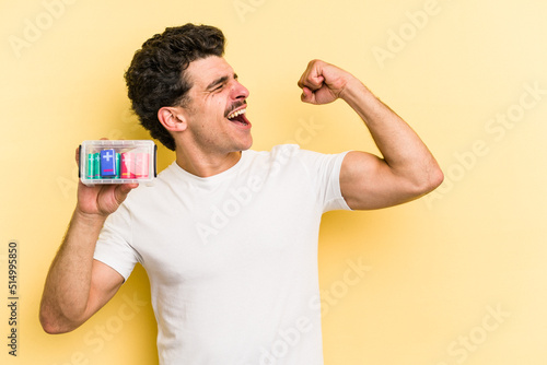 Young caucasian man holding batteries isolated on yellow background raising fist after a victory, winner concept.