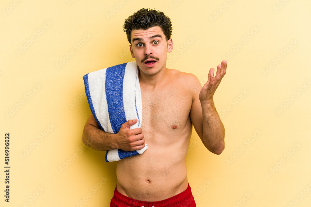 Young caucasian man holding beach towel isolated on yellow background surprised and shocked.