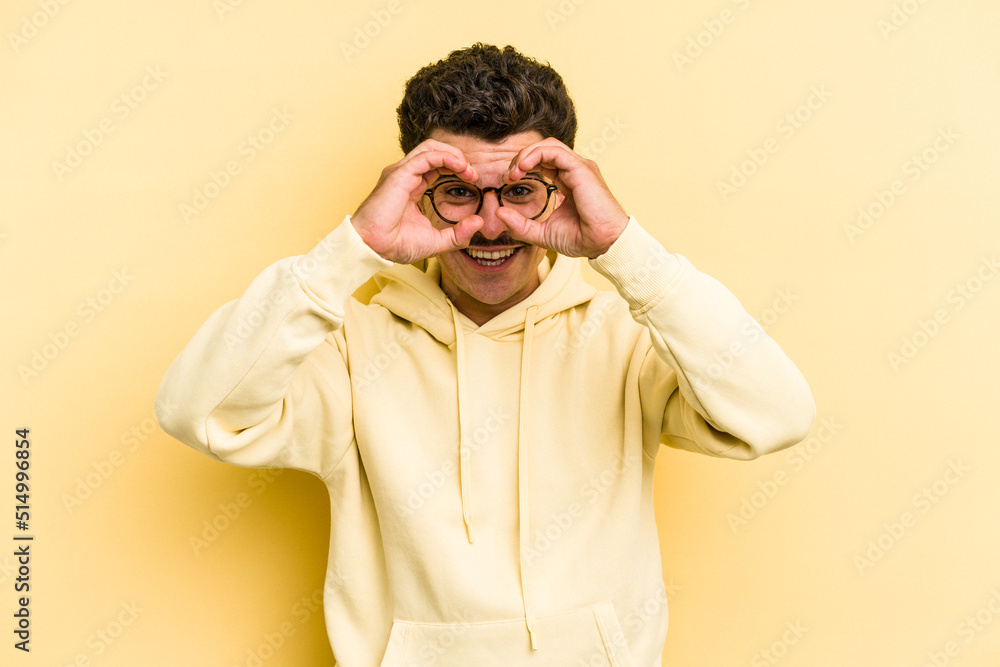 Young caucasian man isolated on yellow background showing okay sign over eyes Young caucasian man isolated on yellow background showing okay sign over eyes