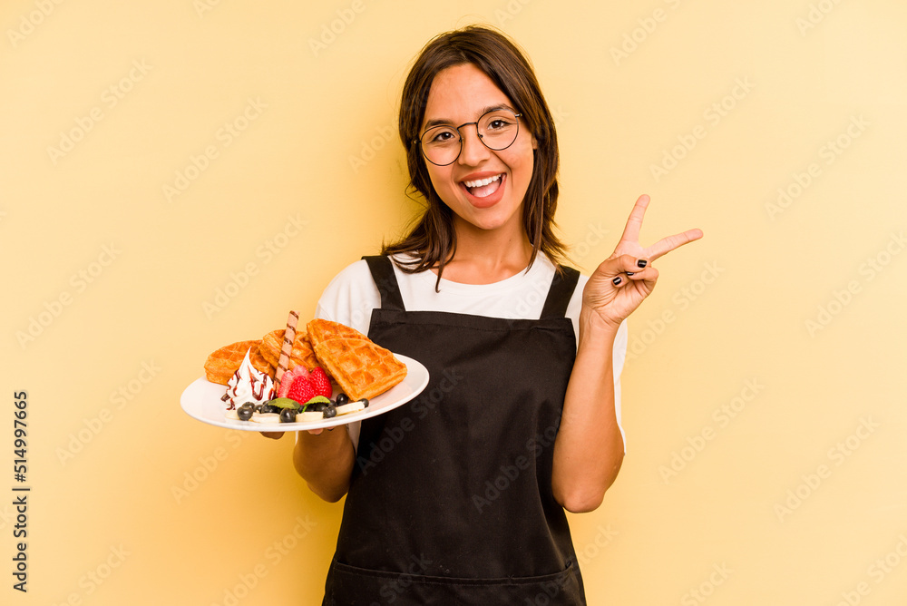 Young hispanic dependent woman holding waffles isolated on yellow background joyful and carefree showing a peace symbol with fingers.