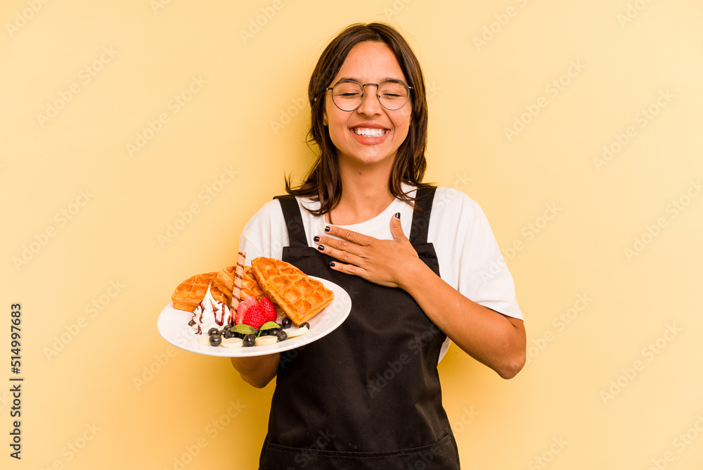 Young hispanic dependent woman holding waffles isolated on yellow background laughs out loudly keeping hand on chest. Young hispanic dependent woman holding waffles isolated on yellow background laughs out loudly keeping hand on chest.