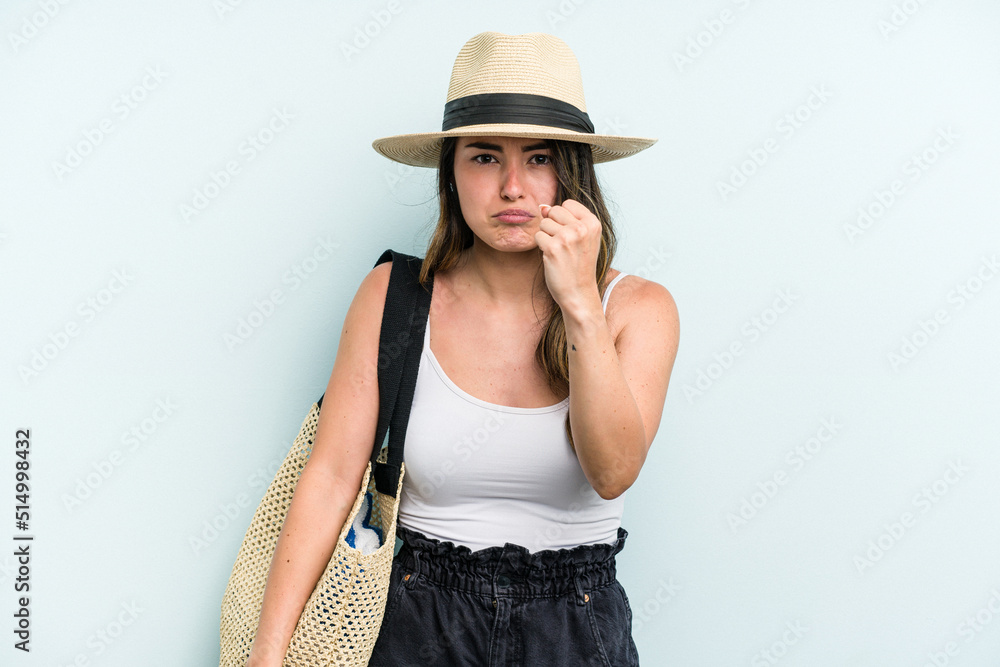Young caucasian woman holding beach bag isolated on blue background showing fist to camera, aggressive facial expression. Young caucasian woman holding beach bag isolated on blue background showing fist to camera, aggressive facial expression.