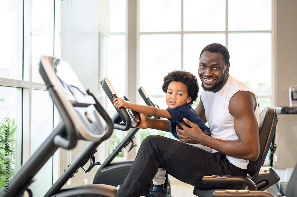 Obraz premium African American man with child exercising on the exercise bike in indoor fitness club. Healthy and Fitness concept.