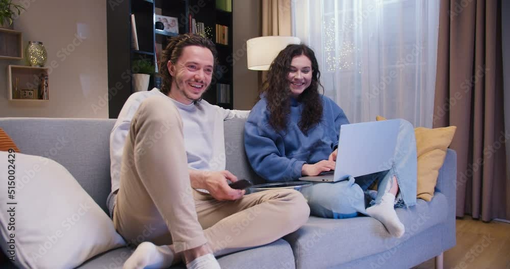 Charming woman working on laptop while her boyfriend watching television at home. Young caucasian couple spending time together and laughing while sitting on sofa in living room. Charming woman working on laptop while her boyfriend watching television at home. Young caucasian couple spending time together and laughing while sitting on sofa in living room.