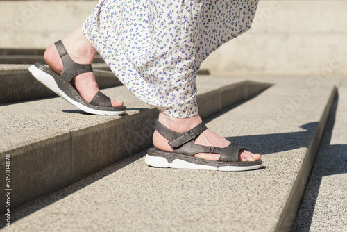 Womens legs in open leather sandals with flat soles go down the stairs. A woman in a light summer dress walks down the stairs.