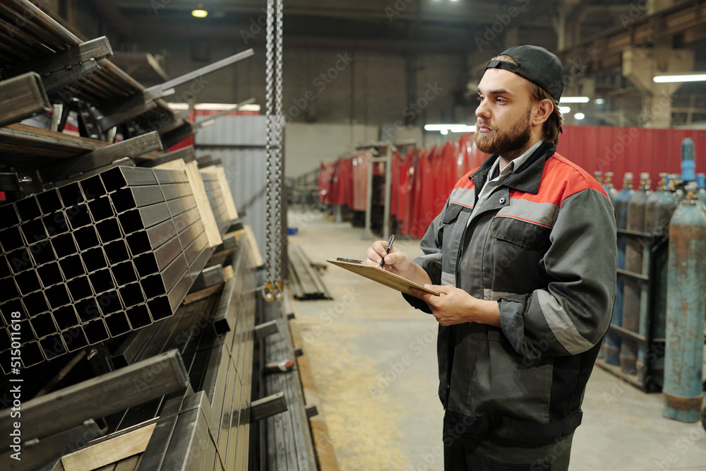 Young engineer in uniform writing down names of work materials for construction of industrial machines put in stacks in workshop